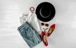 Outfit de mujer con pantalón vaquero, camisa blanca, zapatos de tacón rojo y otros complementos sobre mesa blanca.