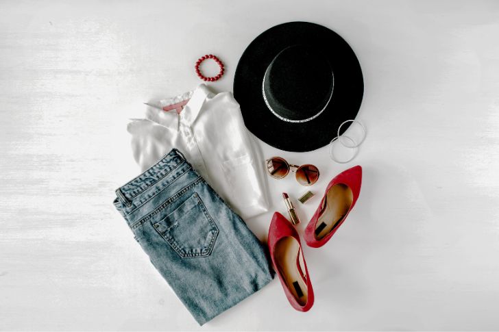 Outfit de mujer con pantalón vaquero, camisa blanca, zapatos de tacón rojo y otros complementos sobre mesa blanca.
