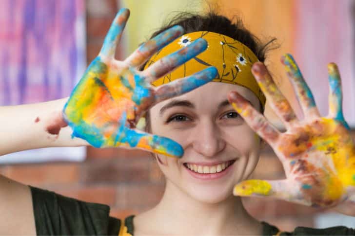 Art Journal. Retrato en primer plano de una joven artista divirtiéndose en un estudio, sonriendo y mostrando las manos sucias de pintura.